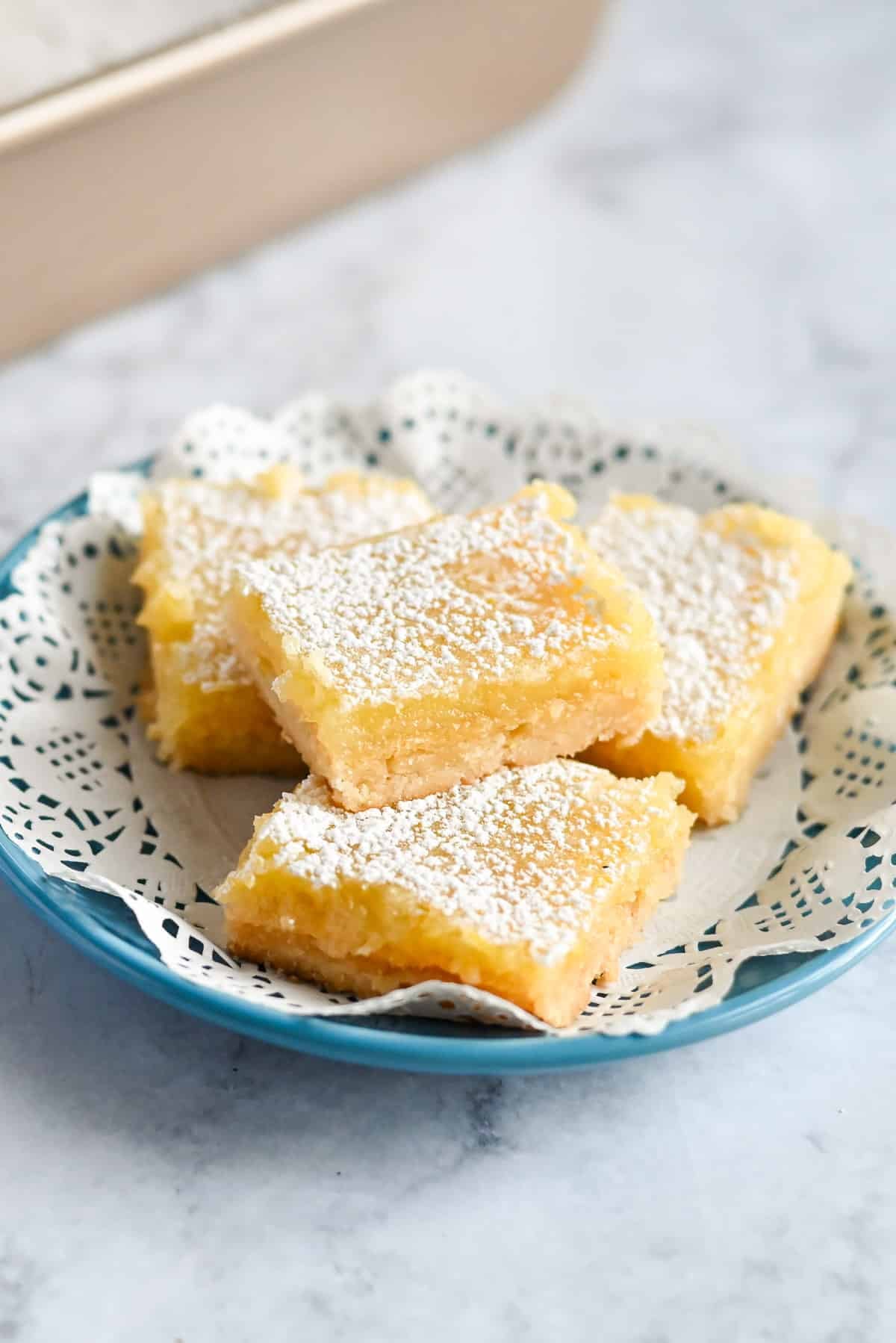Four tart lemon bars dusted with powdered sugar rest on a blue plate with a lace paper doily.