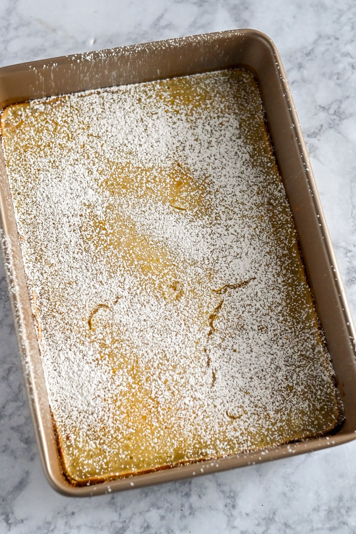 A rectangular baking pan with Tart Lemon Bars topped with powdered sugar on a marble surface.