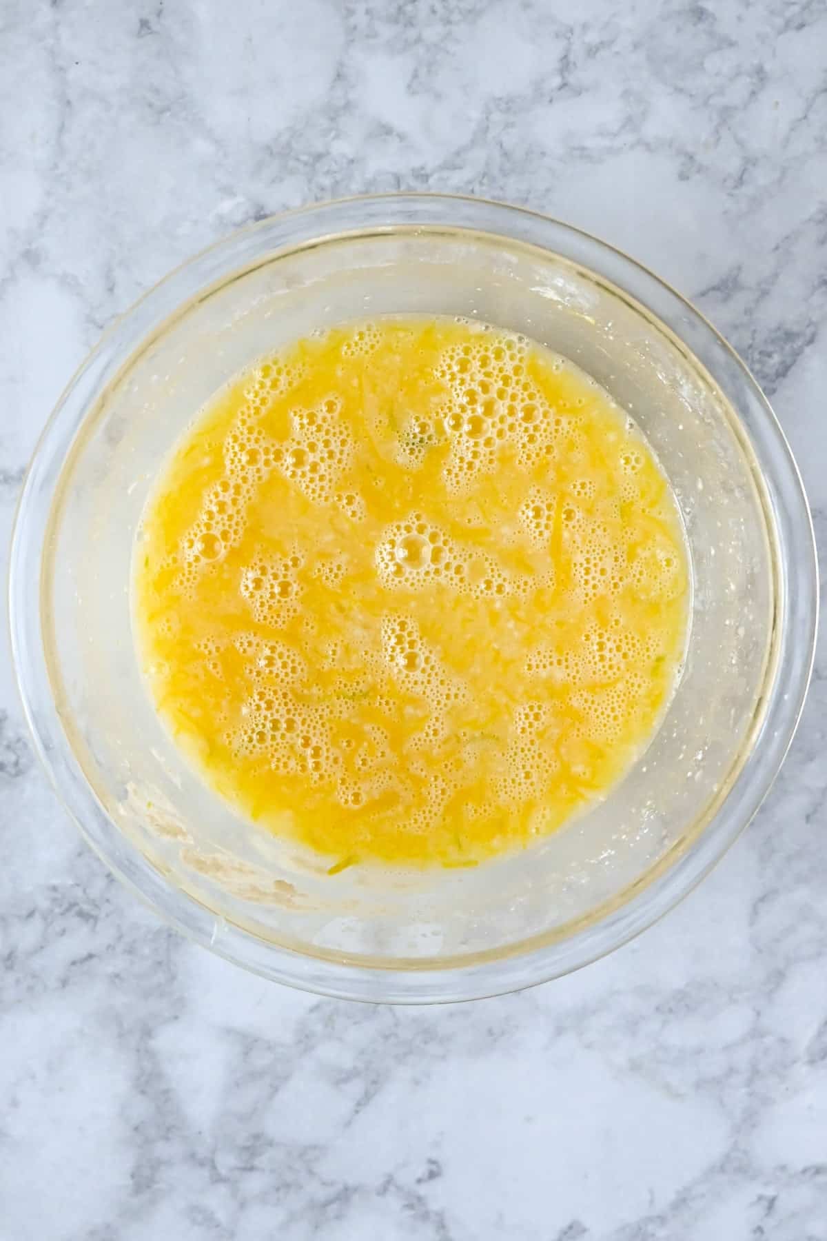 A glass bowl of beaten eggs with bubbles, sitting on a gray marble countertop, ready to be mixed into delicious Tart Lemon Bars.
