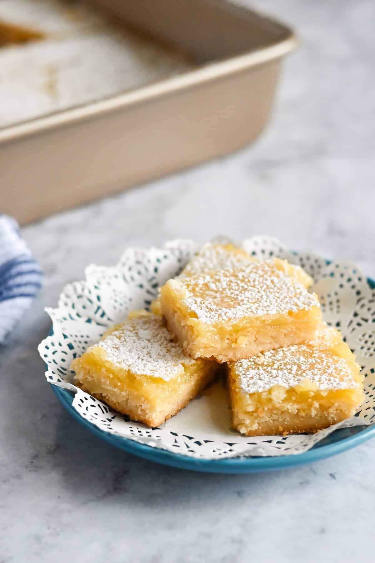 Three Tart Lemon Bars dusted with powdered sugar sit on a blue plate with a white doily, beside a baking pan.