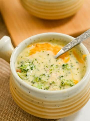 A steaming bowl of creamy broccoli cheese soup sits invitingly on a woven mat, with a spoon ready to dive in.