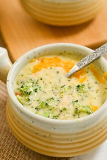 A steaming bowl of creamy broccoli cheese soup sits invitingly on a woven mat, with a spoon ready to dive in.