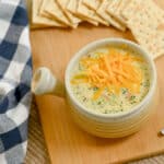 Bowl of creamy Broccoli Cheese Soup topped with shredded cheese, served with crackers on a wooden board.