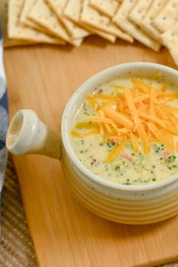 Bowl of creamy Broccoli Cheese Soup topped with shredded cheese, served with crackers on a wooden board.