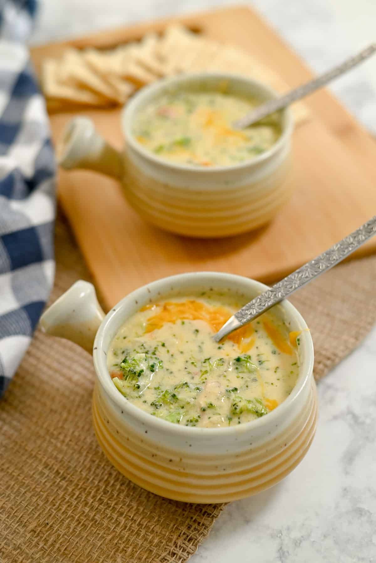 Two bowls of Broccoli Cheese Soup with spoons, served with crackers on a wooden board in the background.