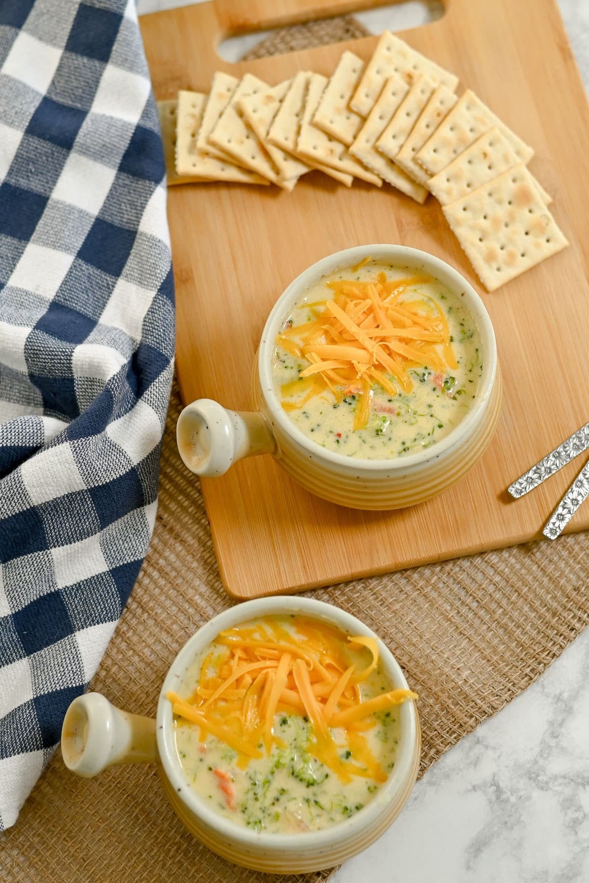 Two bowls of Broccoli Cheese Soup with shredded cheese, served with crackers on a wooden board.