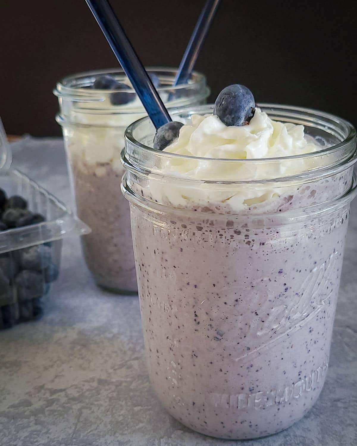 2 mason jars filled with blueberry milkshake next to fresh blueberries.