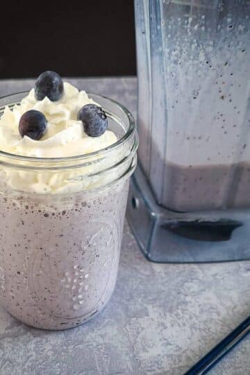 Blueberry milkshake in a mason jar next to fresh berries and a blender.