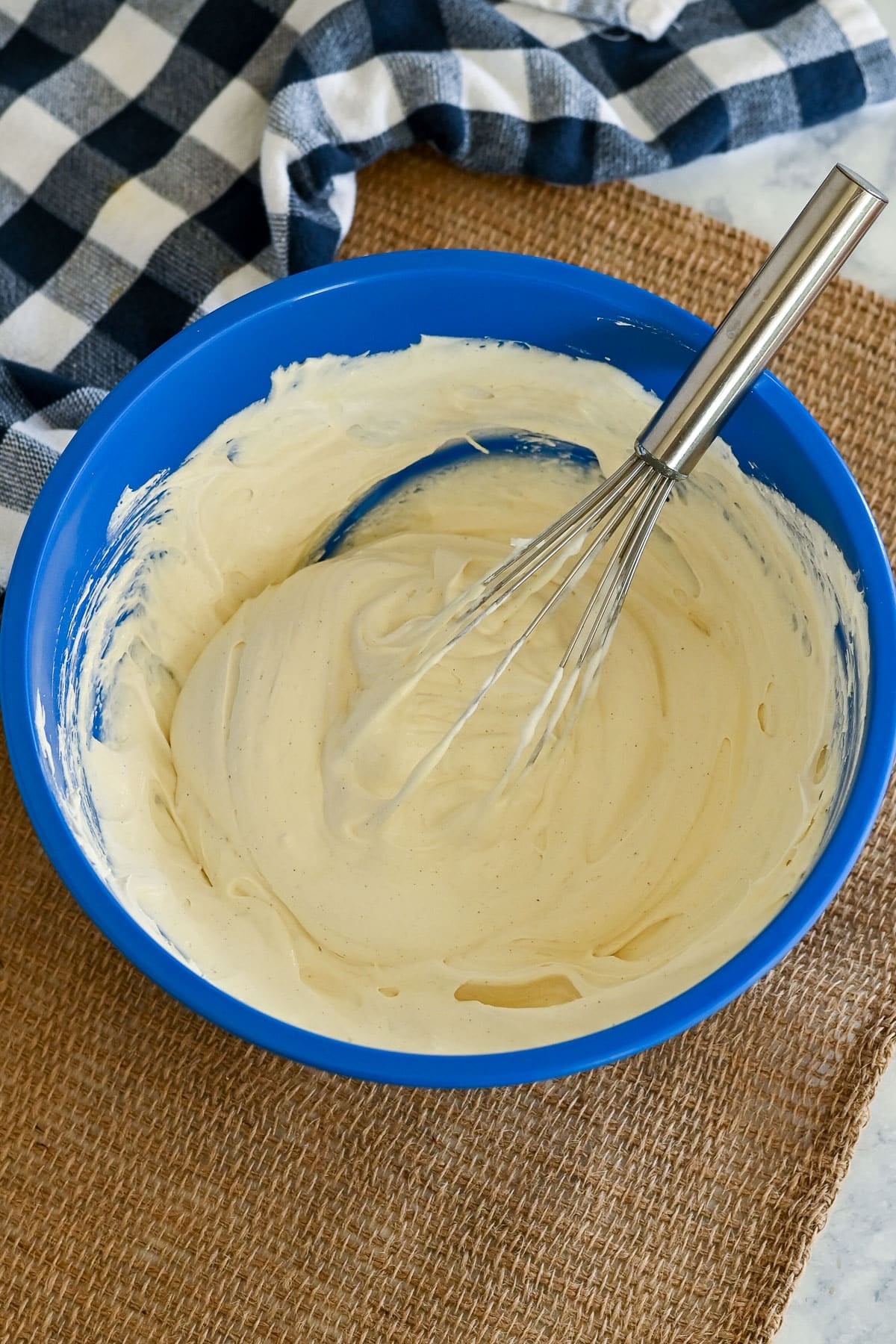 A metal whisk stirs creamy batter for a peaches and cream cake in a blue bowl, set on a burlap mat with a checkered cloth nearby.