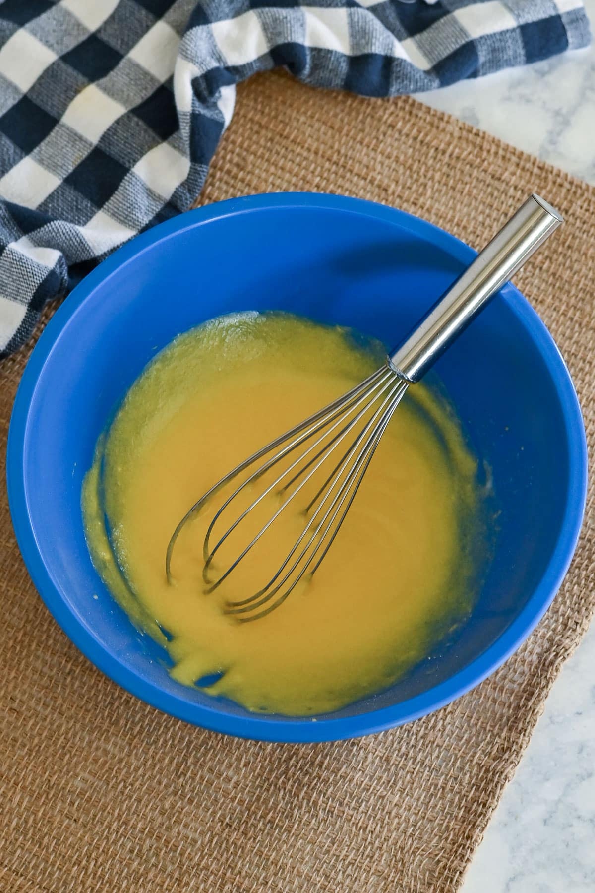 A blue bowl with vanilla pudding for Peaches and Cream Poke Cake sits with a whisk on a burlap mat next to a checkered cloth.