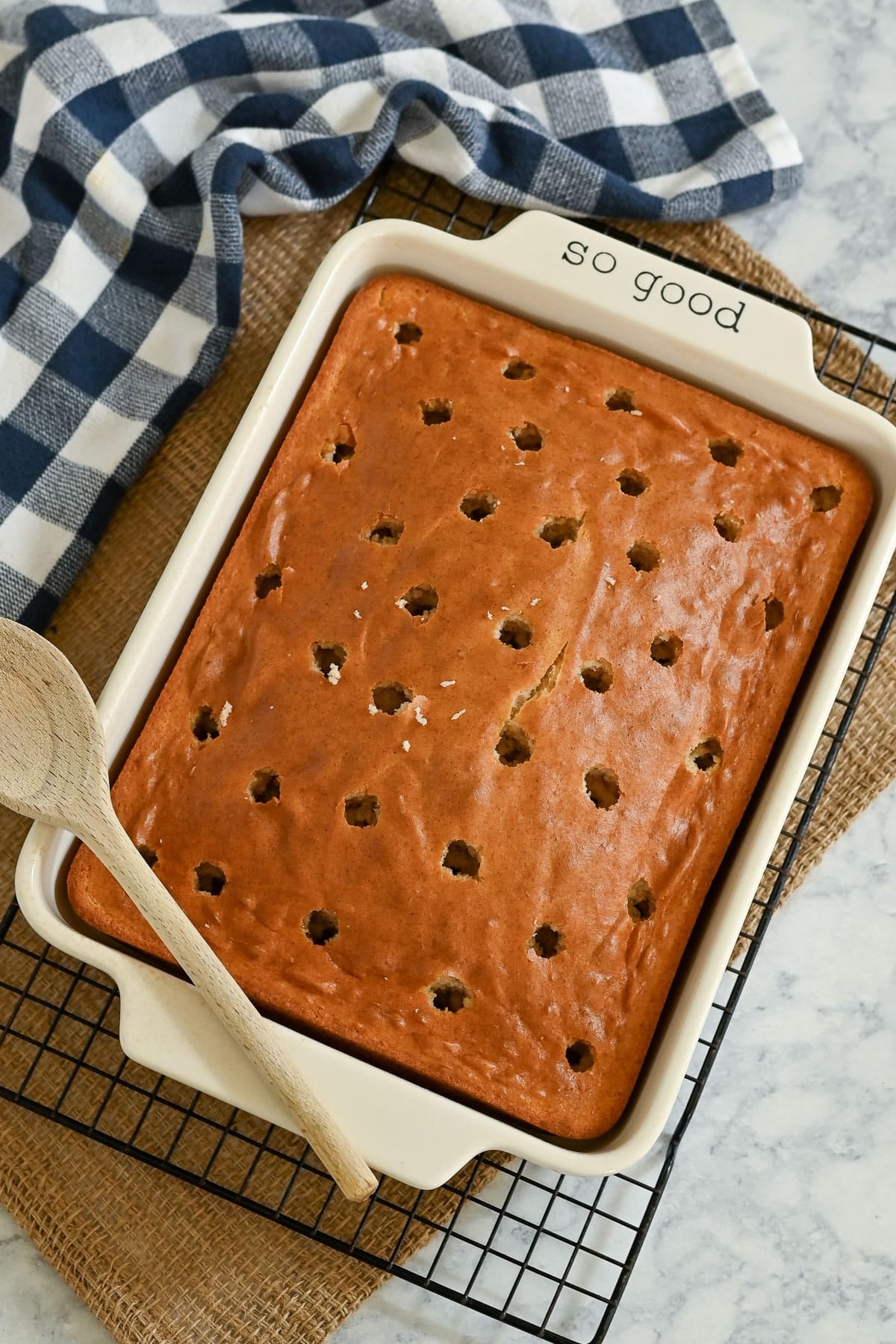 Rectangular Peaches and Cream Poke Cake with holes on top in a baking dish, next to a wooden spoon and checkered towel.