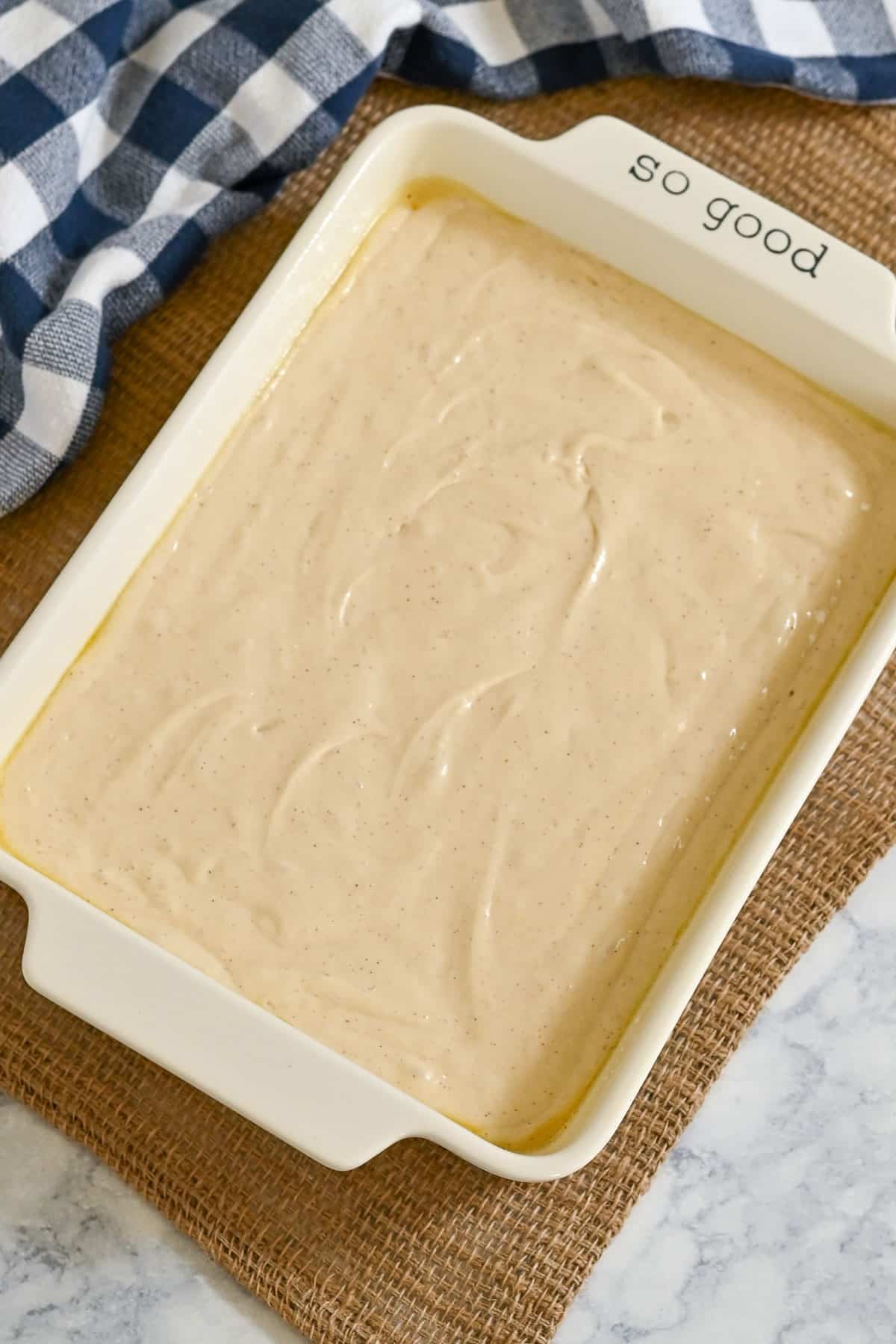 Rectangular dish filled with Peaches and Cream Poke Cake batter, on a burlap mat, next to a blue plaid cloth.
