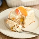 A slice of peaches and cream cake with cream and peach slices sits on a white plate with a fork.