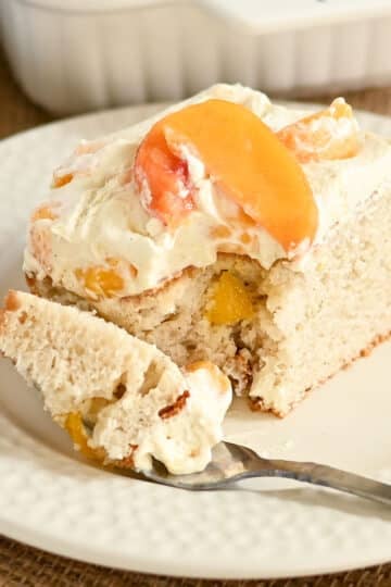 A slice of peaches and cream cake with cream and peach slices sits on a white plate with a fork.