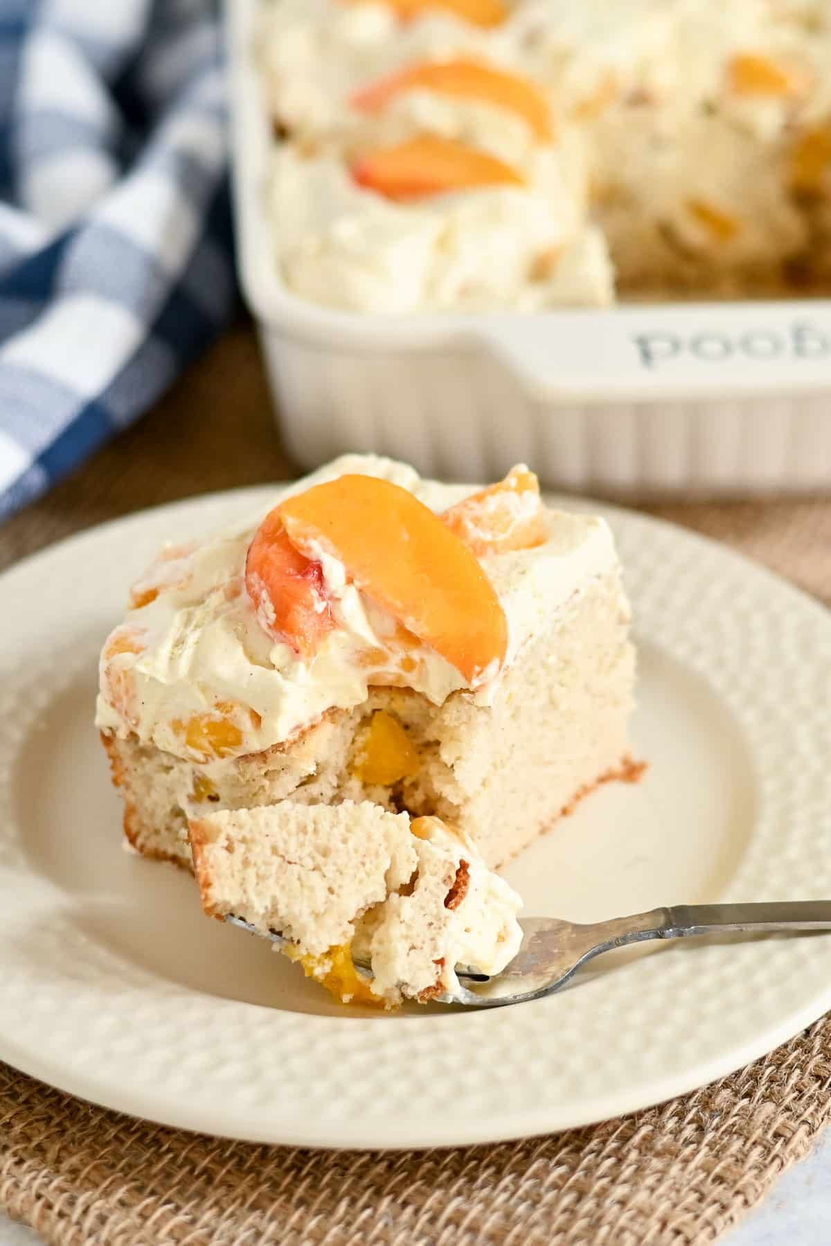 A slice of peaches and cream cake with creamy frosting sits on a plate, a forkful in front, and a baking dish in the background.