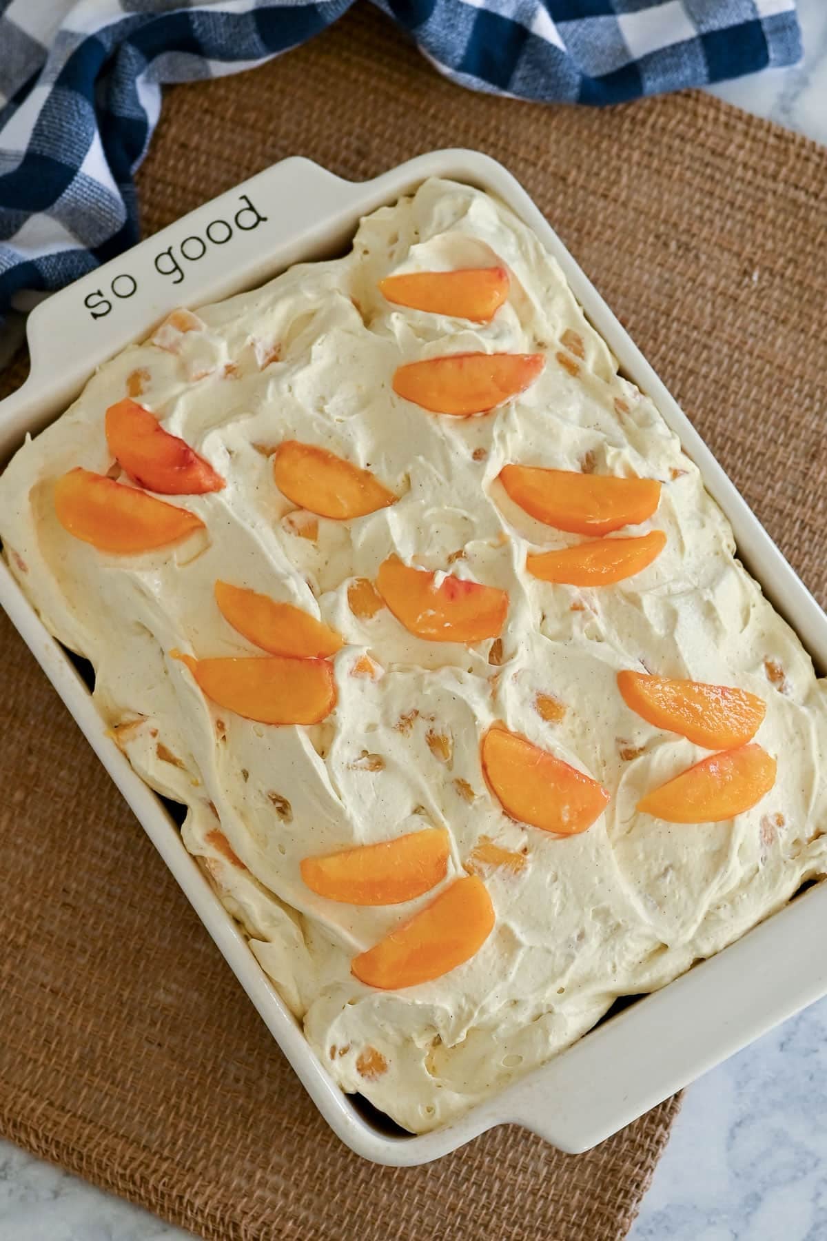 Rectangular dish of creamy peaches and cream cake topped with peach slices, sitting on a woven placemat and blue plaid cloth.