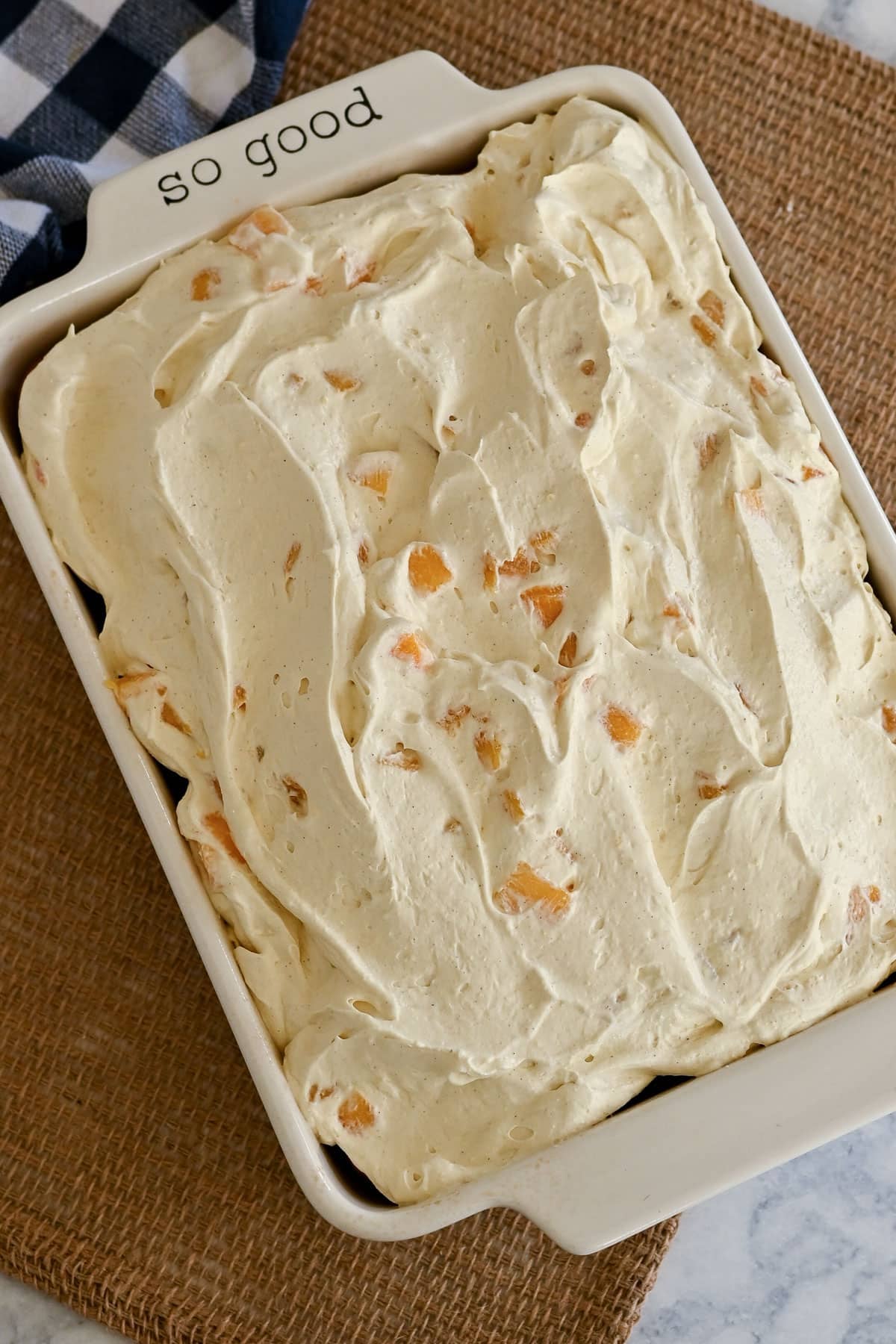 Creamy, nut-studded Peaches and Cream Poke Cake sits in a white casserole dish labeled "so good" on a woven placemat.