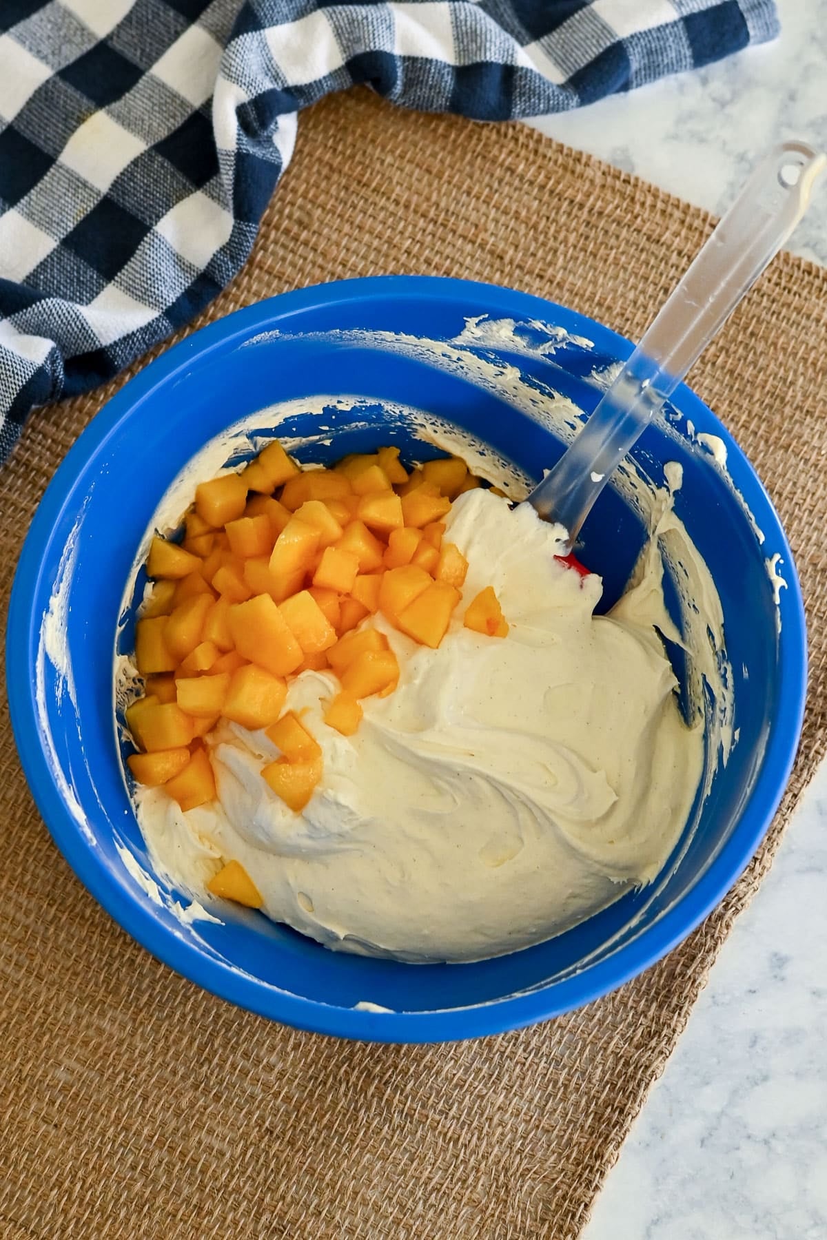 Blue bowl with creamy batter and chopped yellow fruit, perfect for a peaches and cream poke cake, sits next to a checkered cloth on a burlap mat.