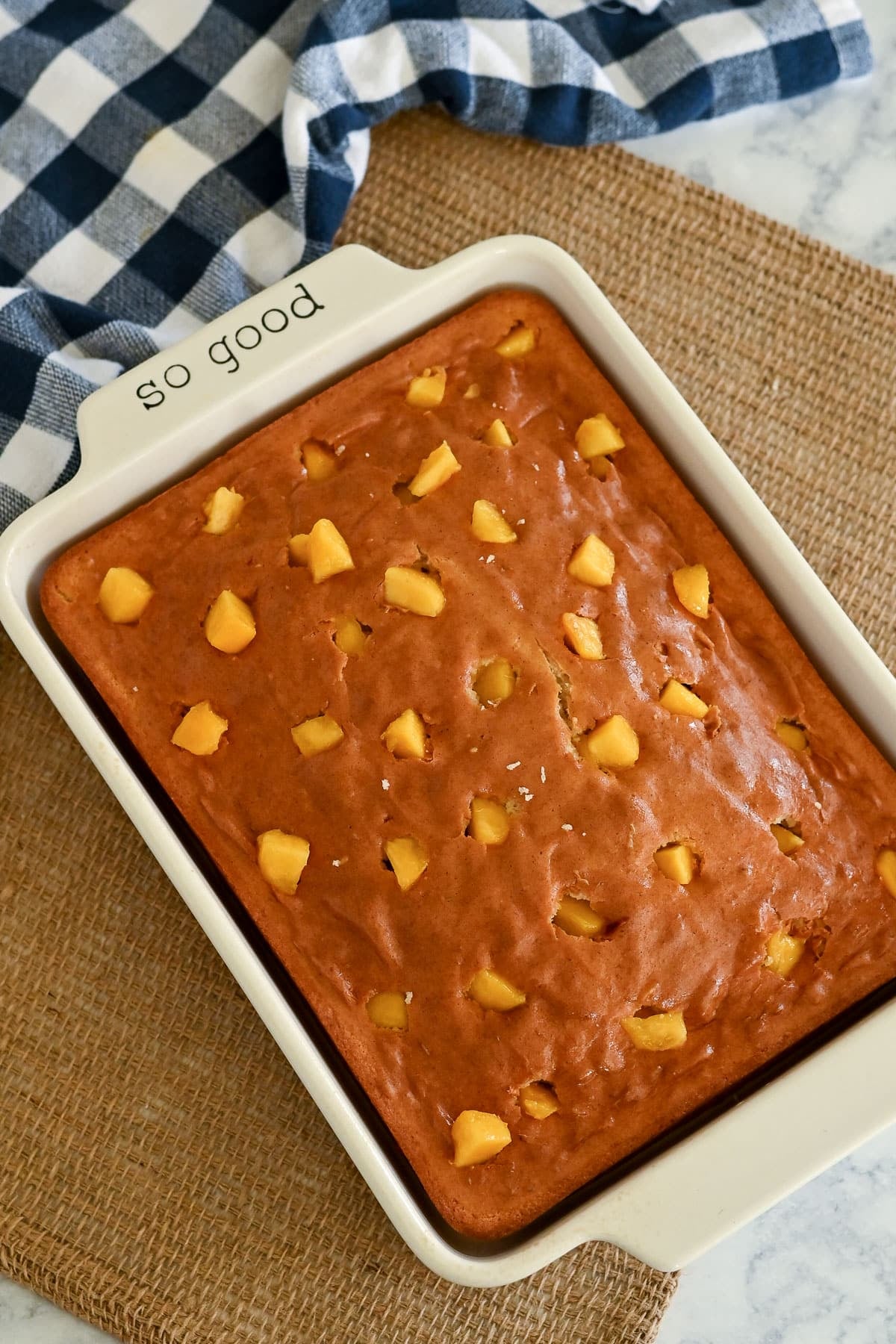 Golden brown Peaches and Cream Poke Cake with fruit chunks in a white baking dish labeled "so good," set atop a burlap mat.
