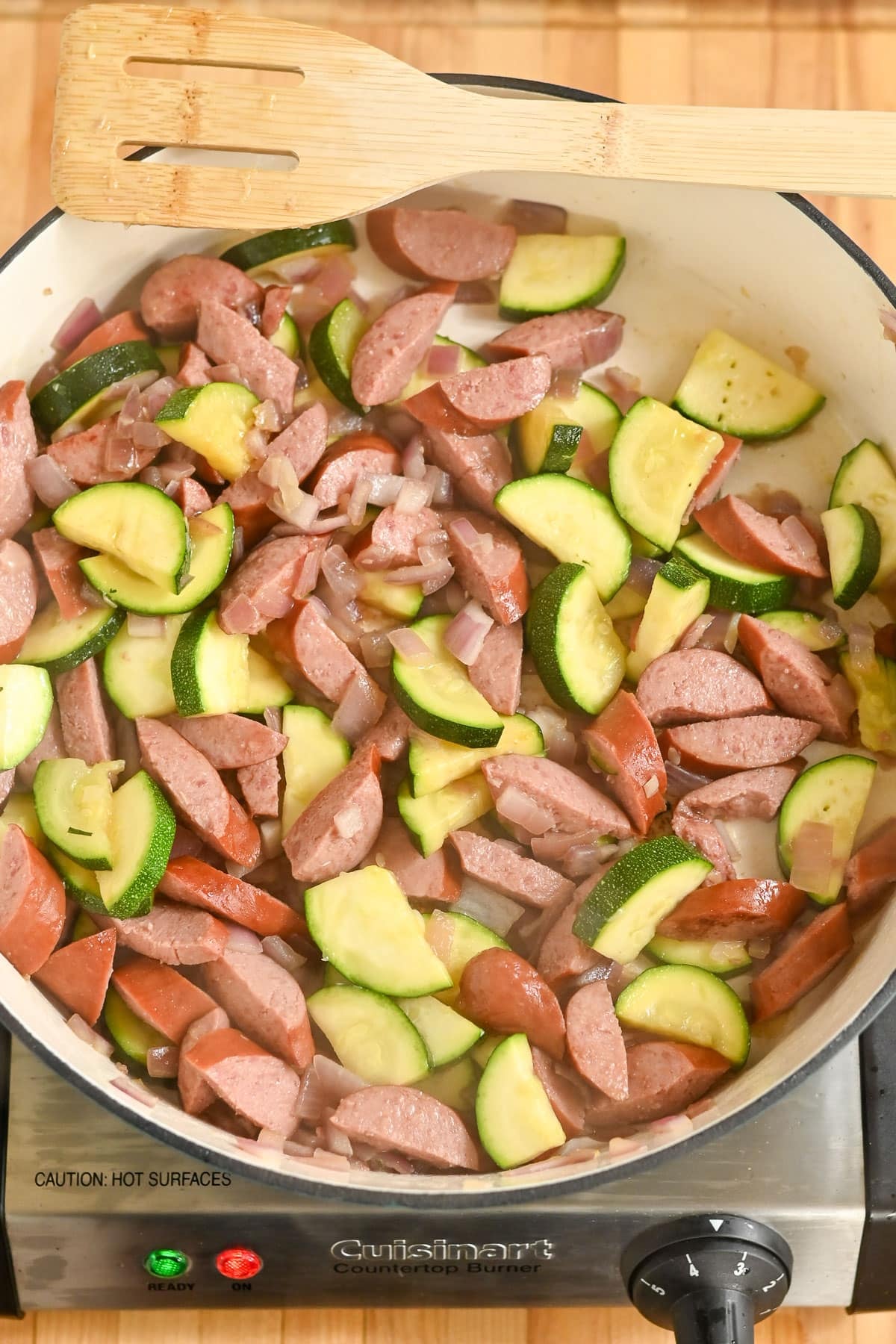 Pasta with Veggies & Smoked Sausage comes together as sliced sausage, zucchini, and onions sizzle in a pan on the stovetop, with a wooden spatula nearby.