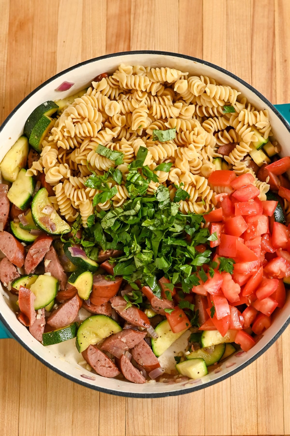 Large skillet with Pasta with Veggies & Smoked Sausage-rotini pasta, sliced sausage, zucchini, tomatoes, and chopped herbs-beautifully arranged on a wooden surface.