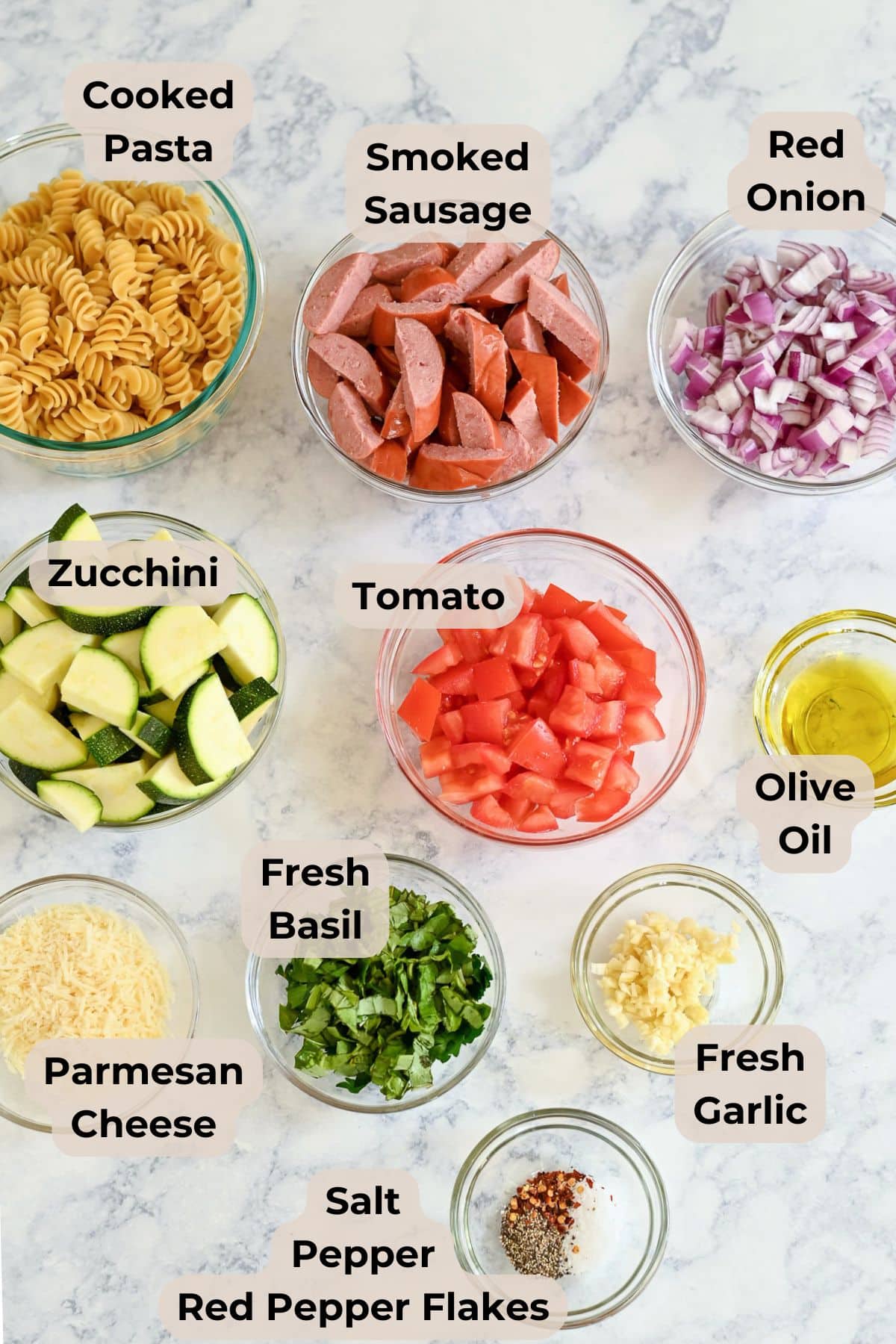 Bowls of Pasta with Veggies & Smoked Sausage, fresh herbs, cheese, and seasonings are arranged on a marble countertop.