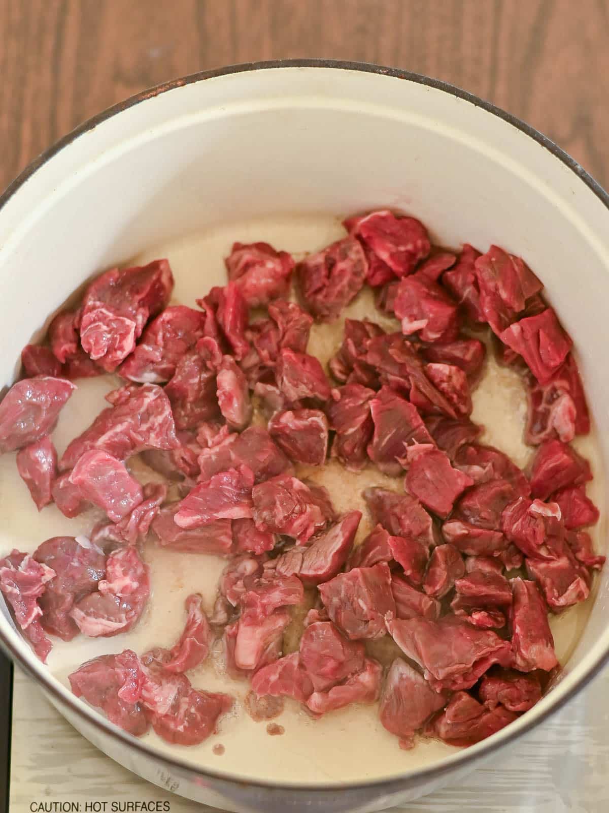 Cubed raw beef for Beef and Dumplings cooking in a white pot on a stovetop, beginning to brown.