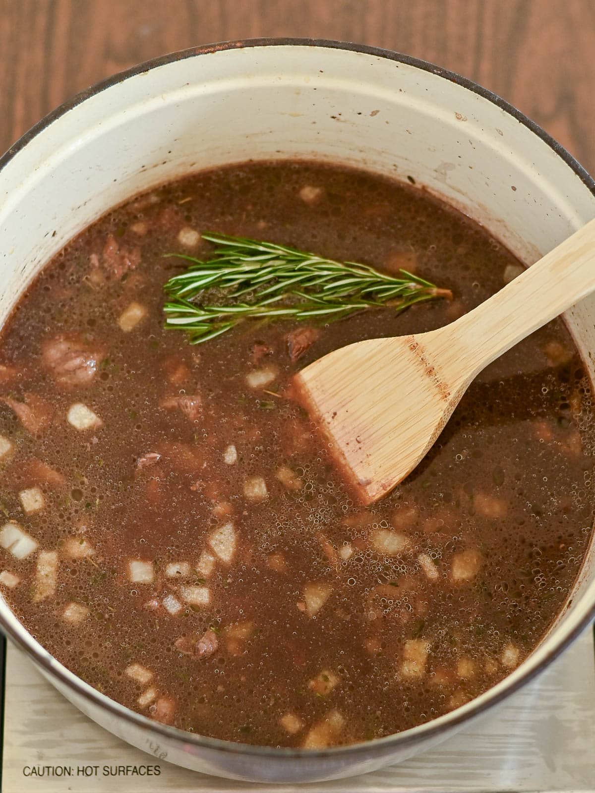 A pot of dark beef and dumplings broth with chopped onions and a sprig of rosemary is being stirred gently with a wooden spatula.