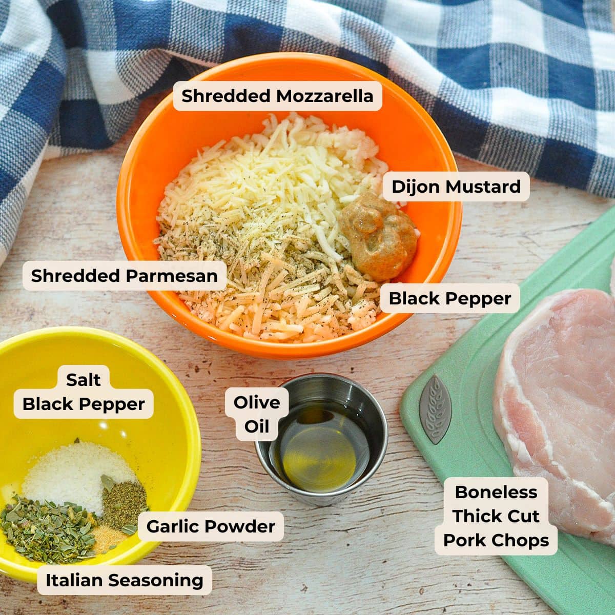 Bowls with shredded cheese, mustard, spices, oil, and thick pork chops on a wooden table with a blue towel-everything you need for easy cheesy pork chops, perfect for a quick dinner.