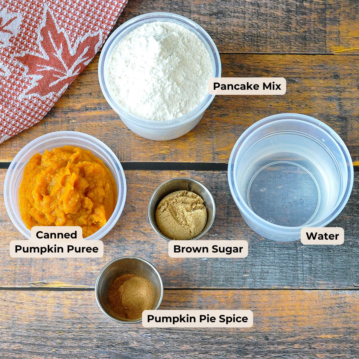 Labeled ingredients for pumpkin pancakes on a wooden table: pancake mix, pumpkin puree, sugar, spice, water.