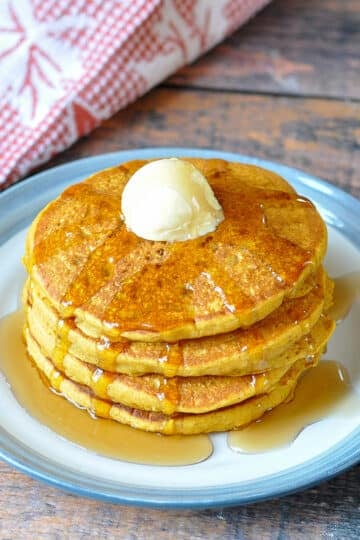 a stack of pumpkin spice pancakes with butter and syrup with a fall themed towel in the back ground