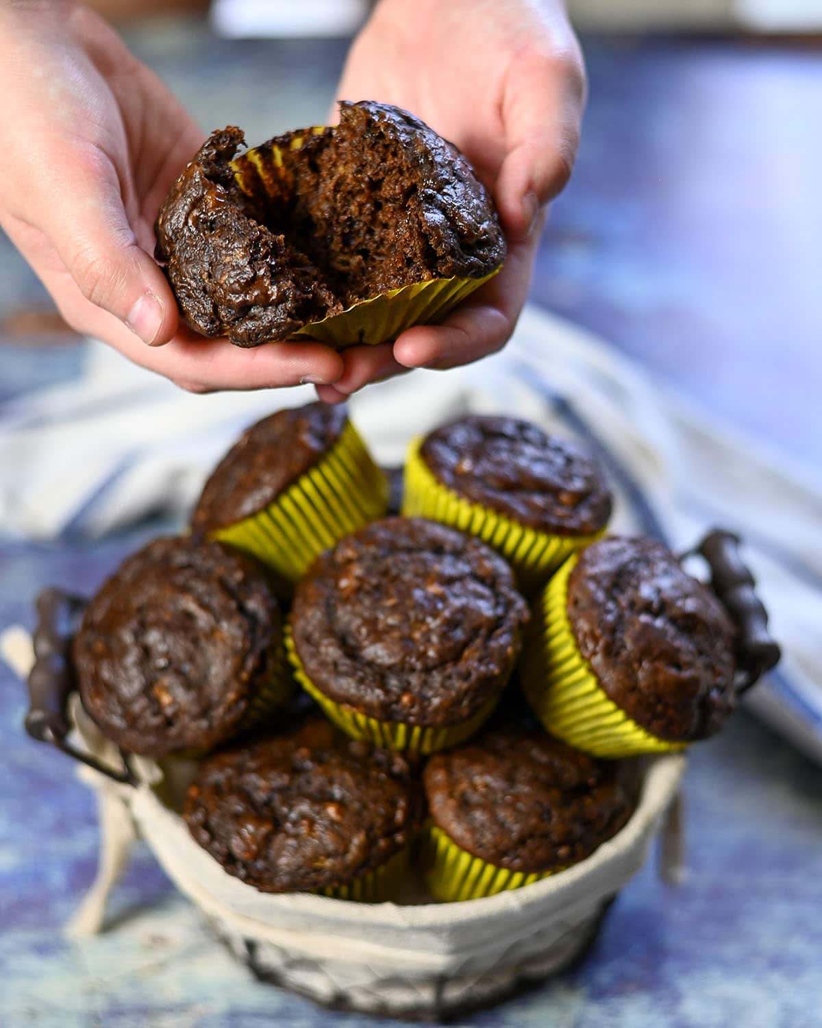 two hands holding a split open chocolate muffin over a wire basket of muffins. 