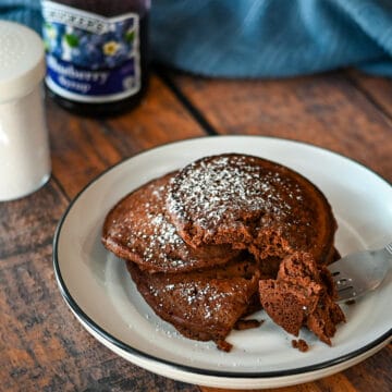 A stack of 3 chocolate pancakes on a white plate next to blueberry syrup.