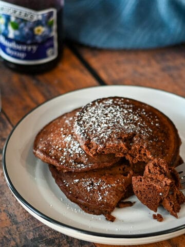 A stack of 3 chocolate pancakes on a white plate next to blueberry syrup.