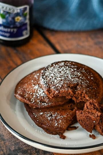 A stack of 3 chocolate pancakes on a white plate next to blueberry syrup.