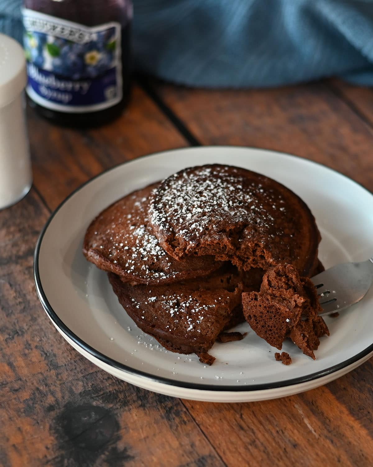 a stack of 3 chocolate pancakes on a white plate next to blueberry syrup. 