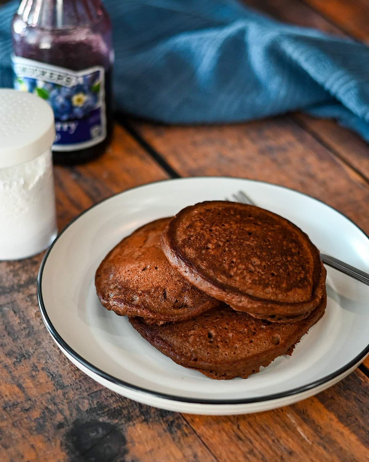 A white plate with chocolate pancakes stacked on it next to powdered sugar and blueberry syrup. 