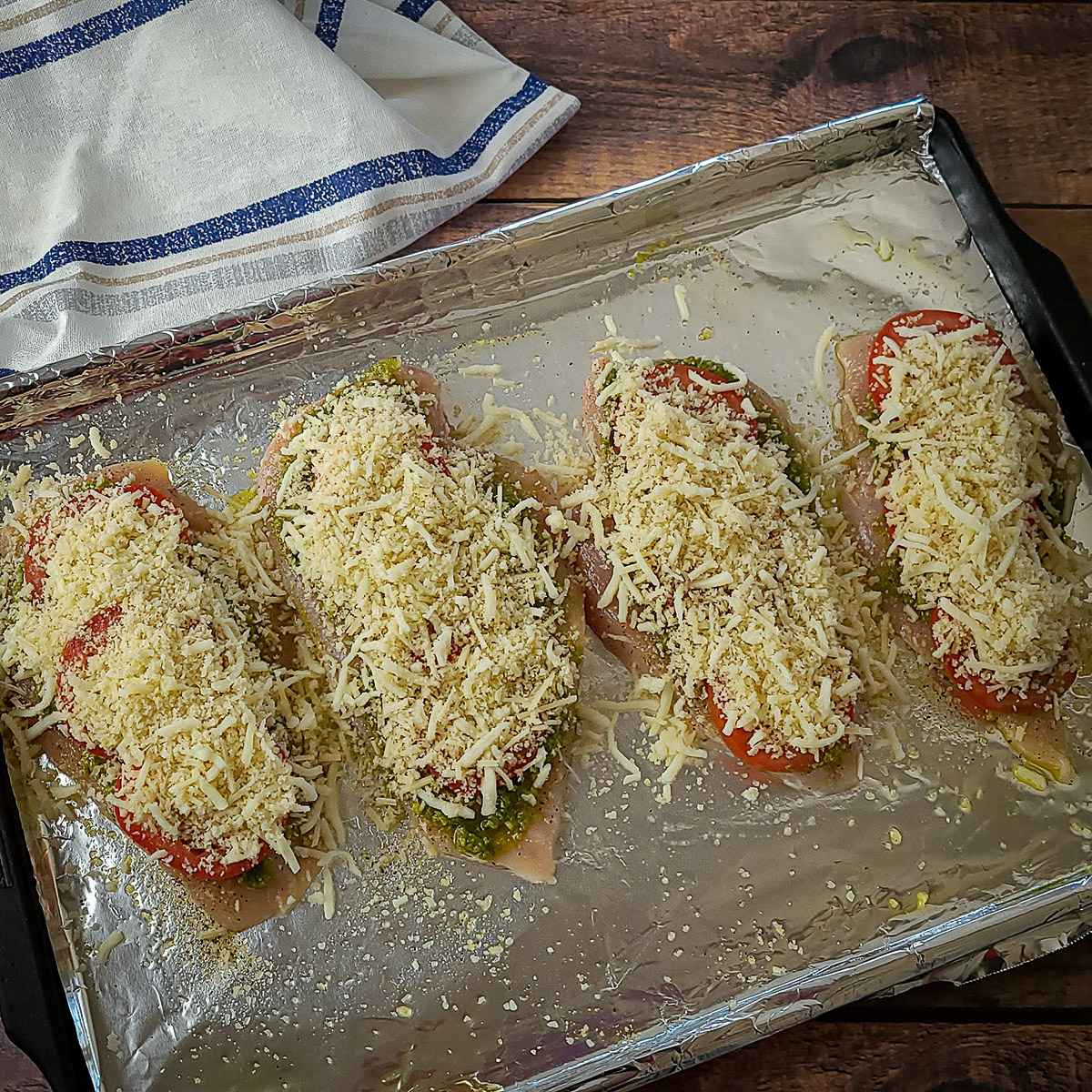 raw chicken covered with tomato and cheese on a foil lined baking sheet. 