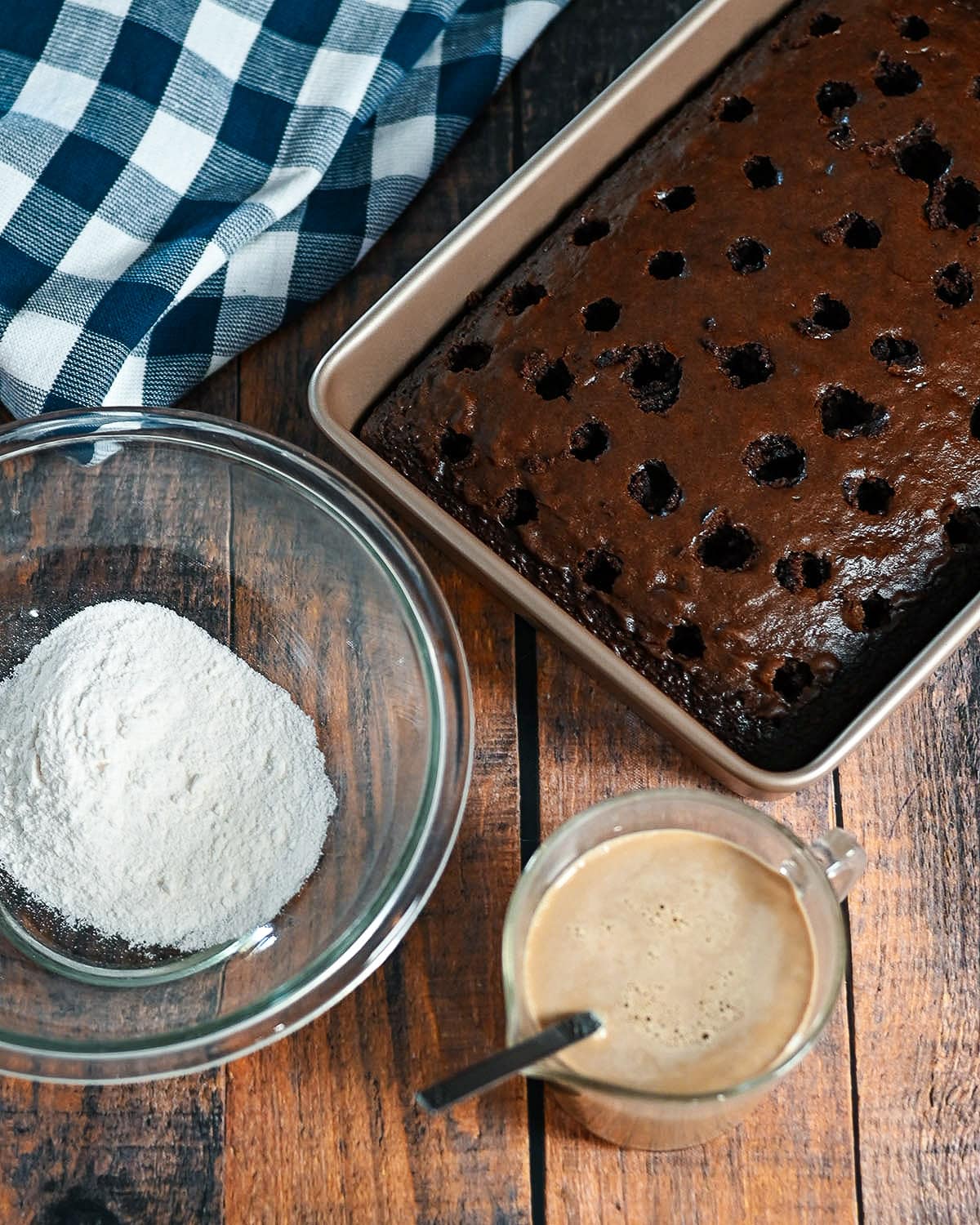 A chocolate cake with holes poked in it next to a bowl and measuring cup. 