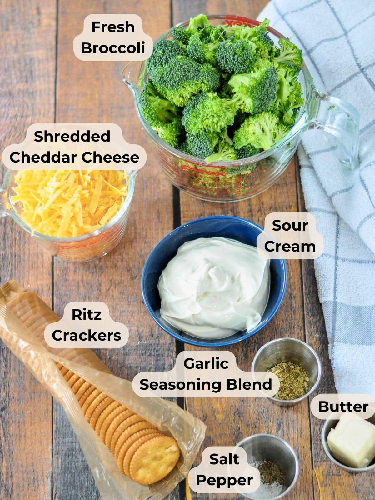Overhead view of ingredients for Simple Broccoli Casserole-broccoli, cheddar cheese, sour cream, Ritz crackers, butter, and seasonings arranged on a wooden table.