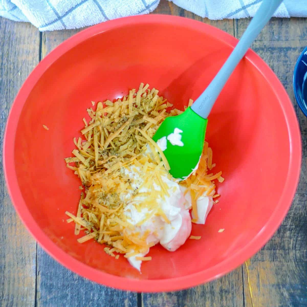 A red bowl with shredded cheese, mayonnaise, and seasonings for Simple Broccoli Casserole is being mixed with a green spatula on a wooden table.