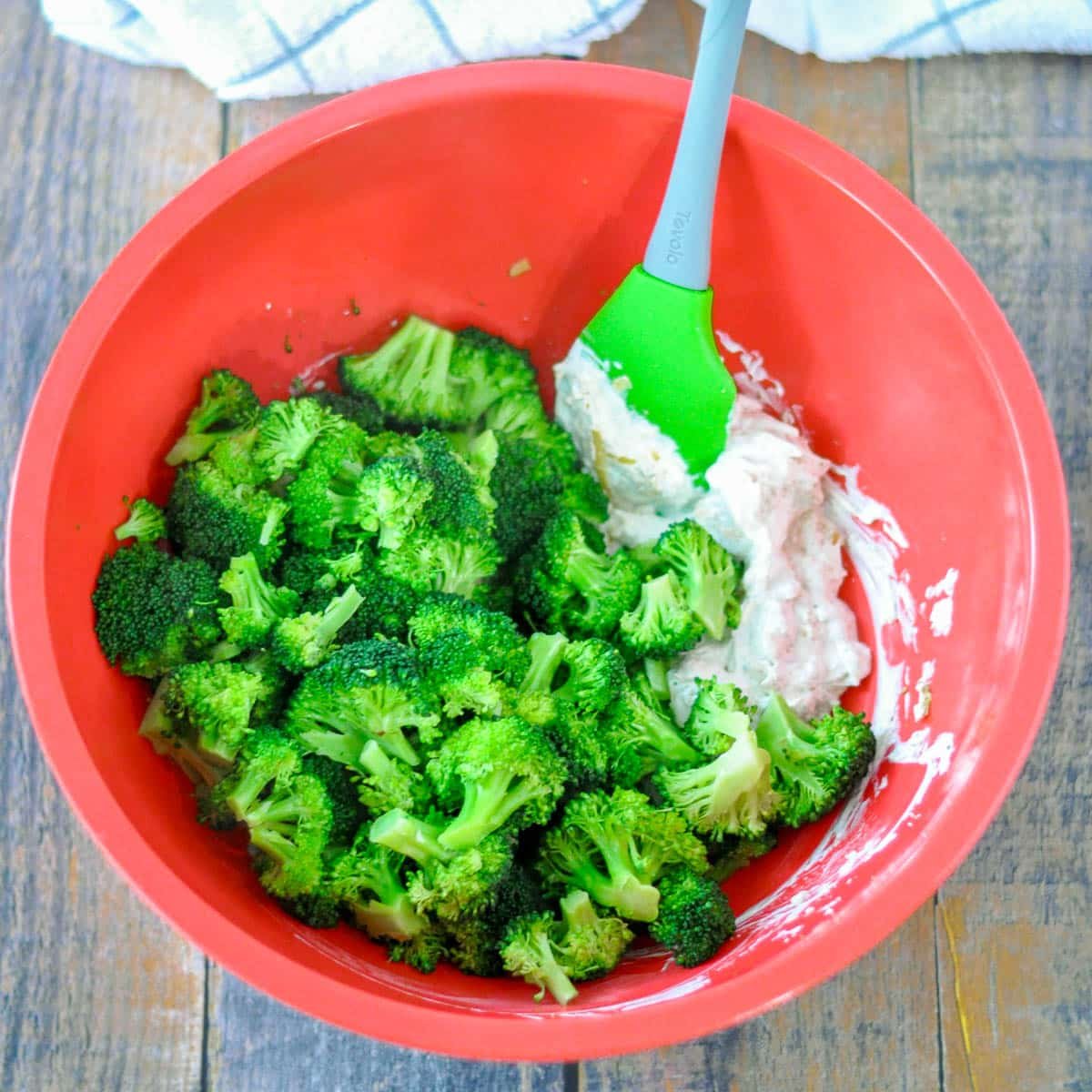 Broccoli florets in a red bowl are being mixed with a creamy white sauce using a green spatula, perfect for an easy Simple Broccoli Casserole.