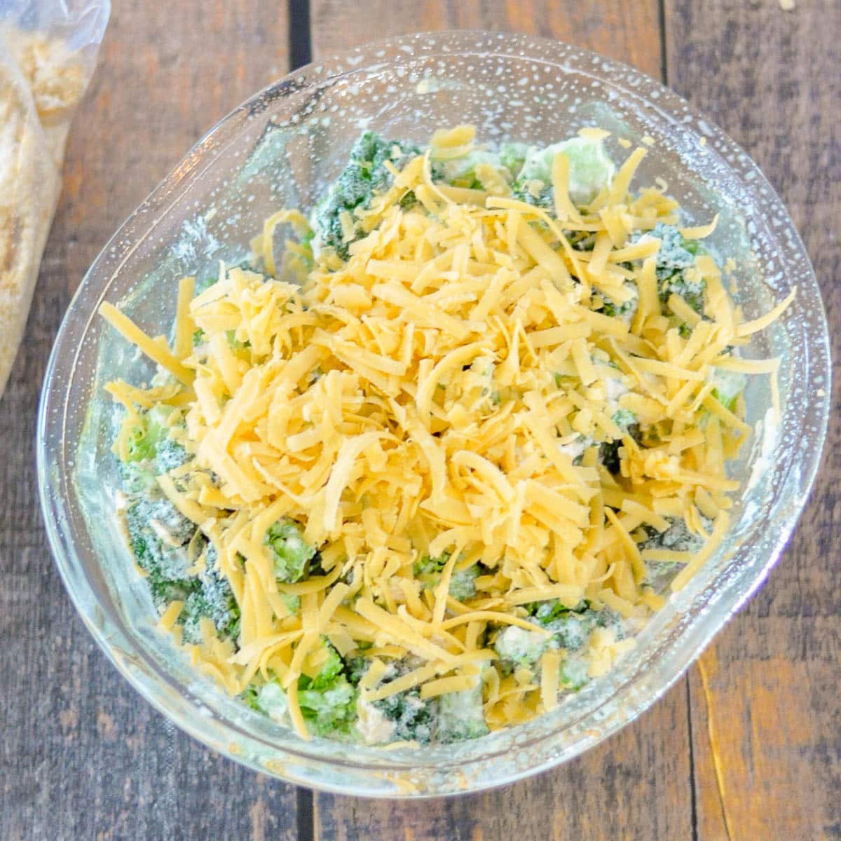 A glass bowl filled with broccoli, shredded cheese, and a creamy mixture sits on a wooden surface, ready to become a Simple Broccoli Casserole.
