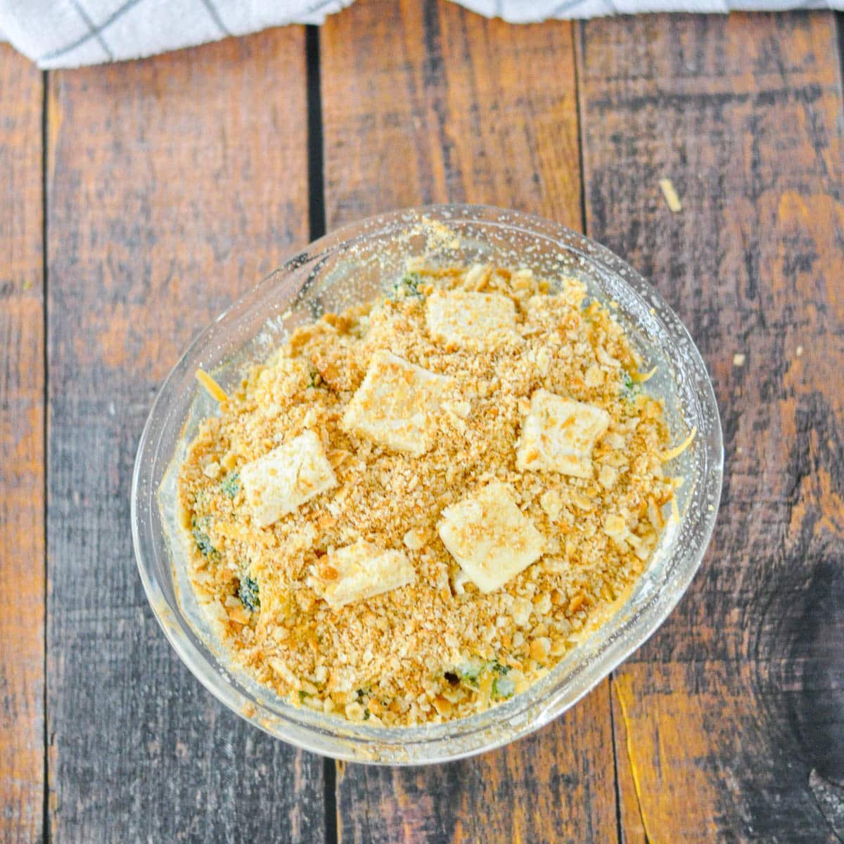 A glass bowl of Simple Broccoli Casserole, topped with golden cracker crumbs, sits on a rustic wooden table.