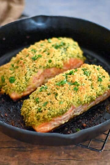 Two salmon filets topped with pesto and panko in a cast iron skillet.