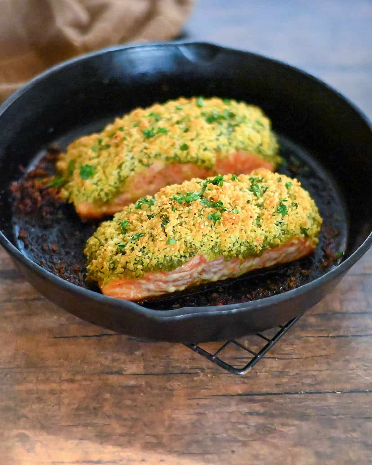 Two salmon filets topped with pesto and panko in a cast iron skillet. 