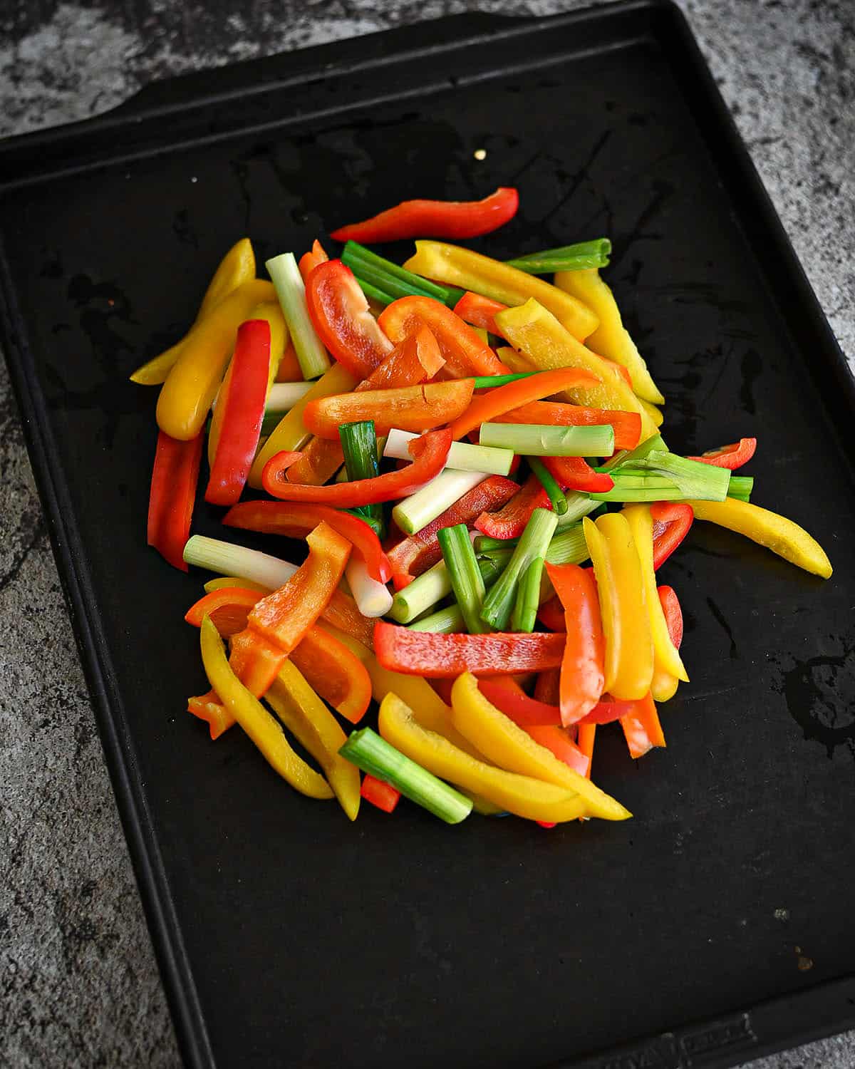 Sliced colorful peppers and green onions on a black baking sheet.
