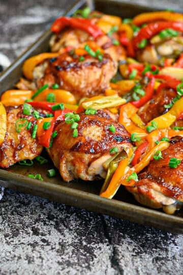 Baked Sweet and Sour Chicken thighs with red and yellow bell peppers, topped with chopped green onions on a baking tray, served with an orange sweet and sour sauce.