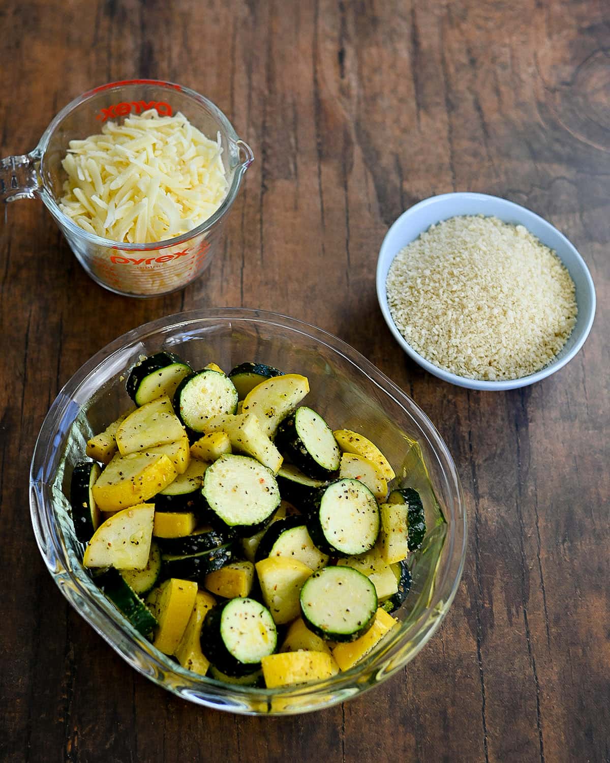 Fresh zucchini and yellow squash in a clear baking dish next to measured cheese and panko.