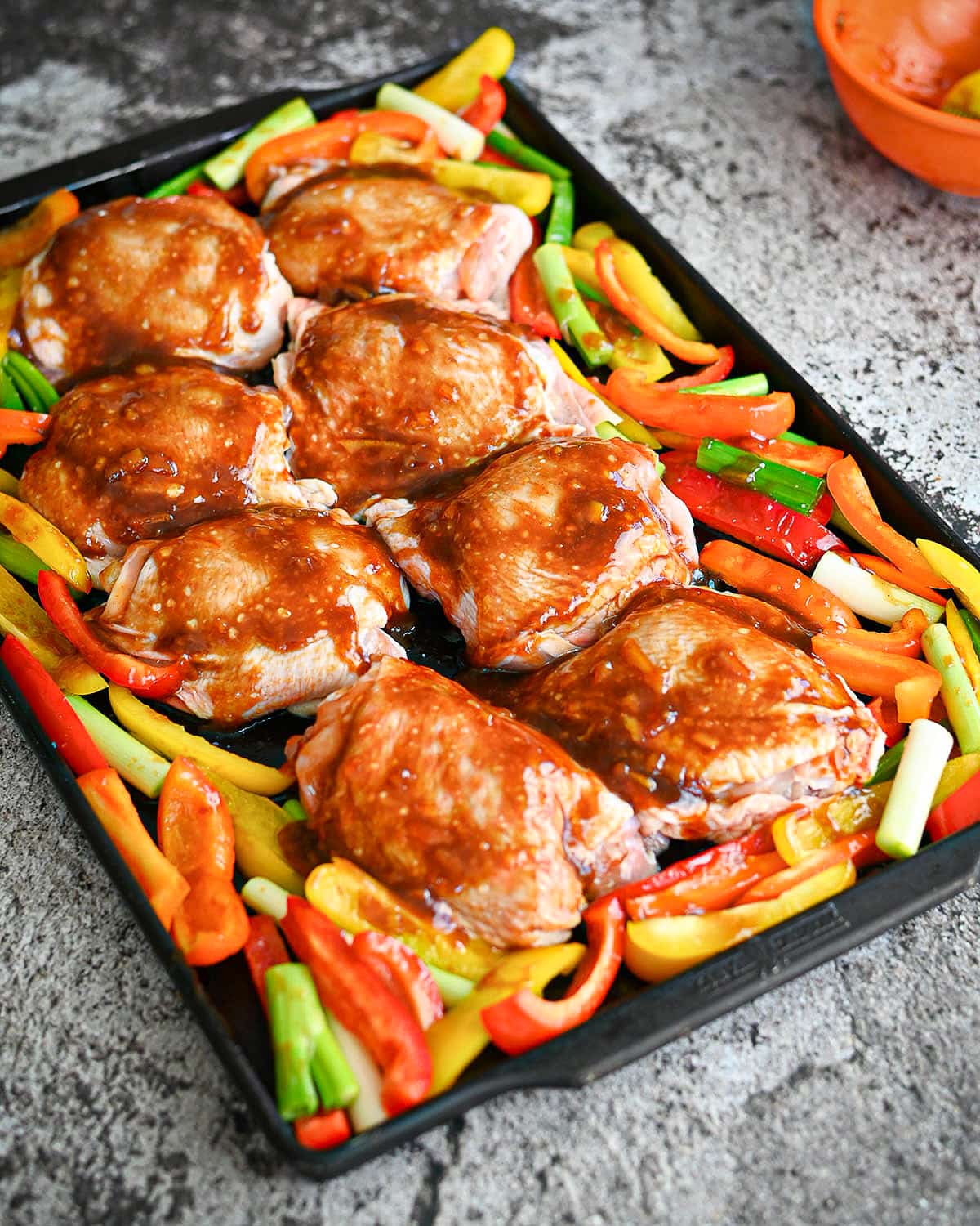 Colorful peppers and raw chicken thighs covered in orange sauce ready to bake. 